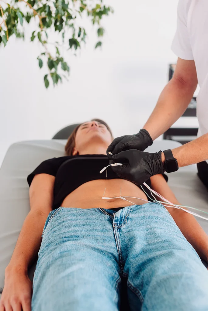 Zabieg elektroakupunktury w Uzdrovisko – terapeuta w czarnych rękawiczkach podłącza przewody do igieł wbitych w brzuch pacjentki.